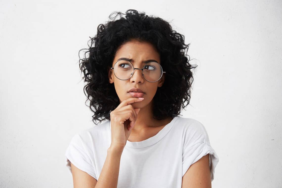 Isolated portrait of stylish young mixed race woman with dark shaggy hair touching her chin and looking sideways with doubtful and sceptical expression, suspecting her boyfriend of lying to her